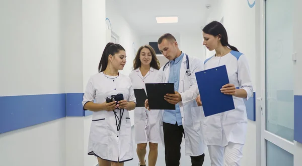 Team of Four Young Medical Praktikanter Diskutera diagnosen av en patient för ytterligare behandling under promenader i en sjukhushall. Praktiskt arbete för praktikanter i vita beläggningar.Läkare på kliniken — Stockfoto