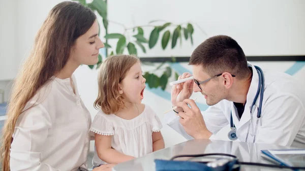 Médico pediatra examinando a una niña triste en visita médica con madre en el hospital. Médico de familia masculino examinando y consultando a la madre y su hijo enfermo. — Foto de Stock