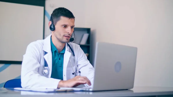 Vänlig manlig läkare i vit medicinsk rock med hörlurar gör konferens samtal på Laptop.Remote Consulting Patient Online från Healthcare Hospital. Telemedicin. — Stockfoto