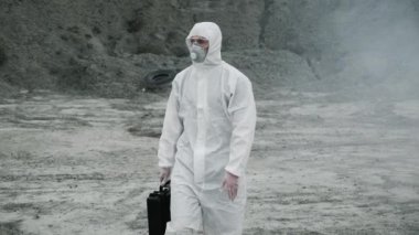 Laboratuvar teknisyeni maske ve kimyasal koruyucu giysi giyip elinde bir alet kutusuyla zehirli dumanın içinden geçiyor.
