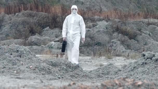 Laboratorní technik v masce a chemickém ochranném obleku, kráčí po suché zemi s krabicí s nářadím přes toxický kouř — Stock video