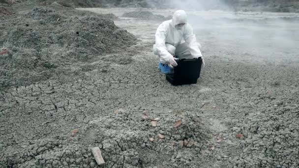 A laboratory technician in a mask and a chemical protective suit opens a toolbox on dry land, around toxic smoke — Stock Video