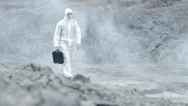 Técnico de laboratório em uma máscara e terno de proteção química, caminha em solo seco com uma caixa de ferramentas através de fumaça tóxica — Vídeo de Stock