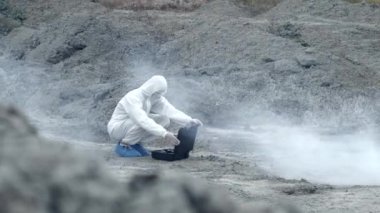 Maskeli bir laboratuar teknisyeni ve kimyasal koruyucu giysi zehirli dumanın etrafındaki kuru arazide bir alet çantası açıyor.