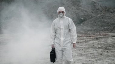 Laboratuvar teknisyeni maske ve kimyasal koruyucu giysi giyip elinde bir alet kutusuyla zehirli dumanın içinden geçiyor.