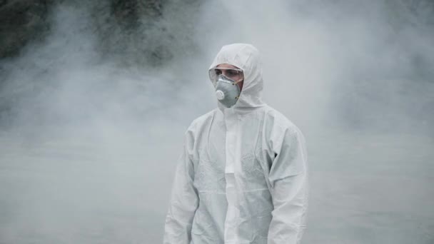 Un tecnico di laboratorio in maschera e una tuta protettiva chimica cammina su terreno asciutto attraverso fumo tossico — Video Stock