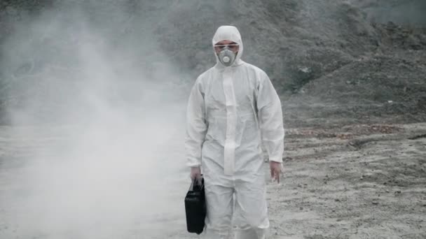 Tecnico di laboratorio in maschera e tuta protettiva chimica, cammina su terreno asciutto con una cassetta degli attrezzi attraverso fumo tossico — Video Stock