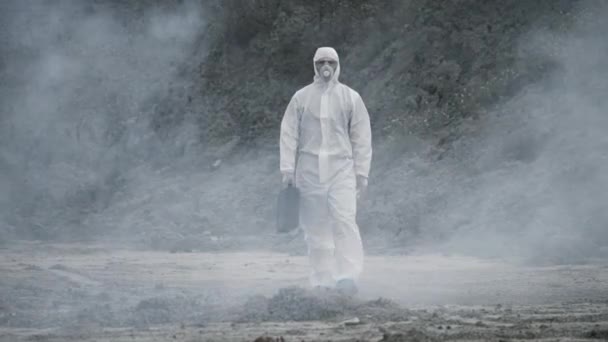 Tecnico di laboratorio in maschera e tuta protettiva chimica, cammina su terreno asciutto con una cassetta degli attrezzi attraverso fumo tossico — Video Stock