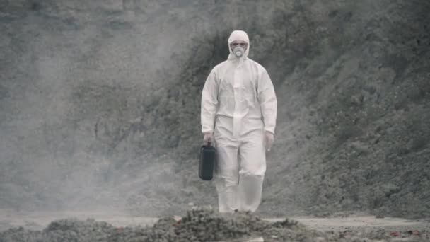 身穿防毒面具和化学防护服的实验室技术员，带着工具箱走在干燥的地面上，穿过有毒的烟雾 — 图库视频影像