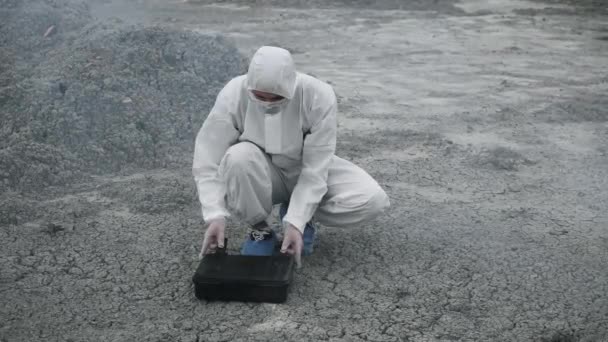 Ein Laborant in Maske und chemischem Schutzanzug öffnet auf dem Trockenen eine Werkzeugkiste um giftigen Rauch herum — Stockvideo