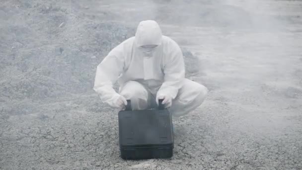 A laboratory technician in a mask and a chemical protective suit opens a toolbox on dry land, around toxic smoke — Stock Video