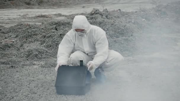 Un técnico de laboratorio con una máscara y un traje de protección química abre una caja de herramientas en tierra firme, alrededor del humo tóxico — Vídeo de stock