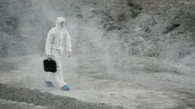 Laboratuvar teknisyeni maske ve kimyasal koruyucu giysi giyip elinde bir alet kutusuyla zehirli dumanın içinden geçiyor.