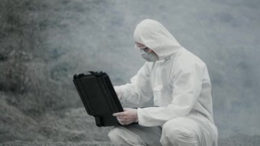 Maskeli bir laboratuar teknisyeni ve kimyasal koruyucu giysi zehirli dumanın etrafındaki kuru arazide bir alet çantası açıyor.