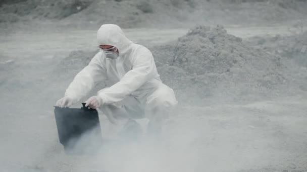 En laboratorietekniker i mask och en kemisk skyddsdräkt öppnar en verktygslåda på torra land, runt giftig rök — Stockvideo