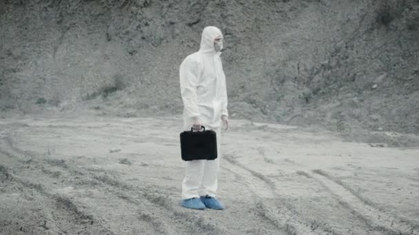 Técnico de laboratorio en una máscara y traje de protección química, camina sobre tierra seca con una caja de herramientas a través de humo tóxico — Vídeos de Stock