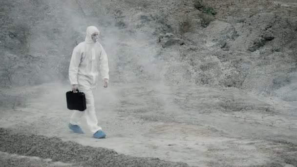 身穿防毒面具和化学防护服的实验室技术员，带着工具箱走在干燥的地面上，穿过有毒的烟雾 — 图库视频影像