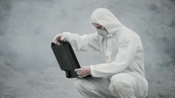 A laboratory technician in a mask and a chemical protective suit opens a toolbox on dry land, around toxic smoke — Stock Video