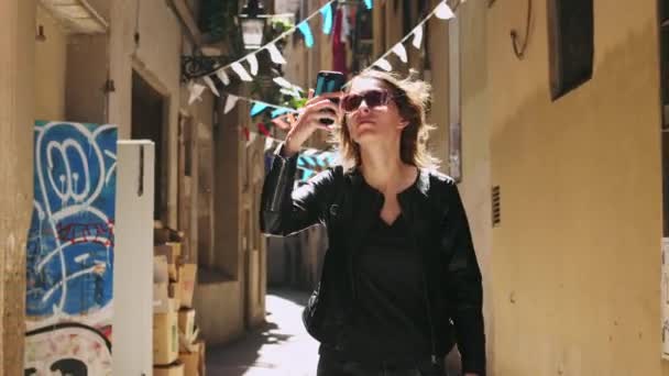Привлекательная туристическая женщина делает фото использует смартфон во время прогулки по узкой улице. — стоковое видео
