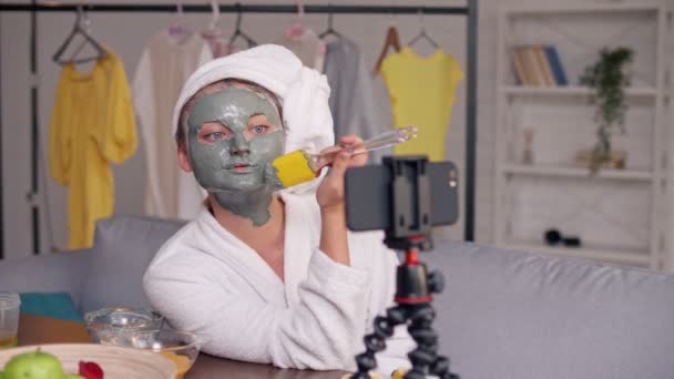 Un Beauty Blogger in un camice bianco con un asciugamano bianco sulla testa Applica una maschera idratante verde al suo viso — Video Stock