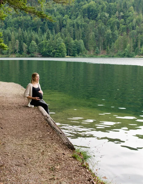 Όμορφη, χαριτωμένη έγκυος γυναίκα κάθεται κοντά σε μια λίμνη στο βουνό σε ένα δάσος στην ακτή — Φωτογραφία Αρχείου