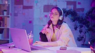 Gen Z Yaşam Tarzı, Dijital Çoklu Görev, Neon Mood. Genç Kız Müzik Dinlerken ve Dizüstü bilgisayar izlerken resim çizer. Neon Işığında Modern Gençliğin Yaratıcı Kaosu 'nda Kişisel İfade