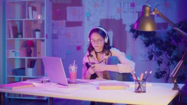 Gen Z Yaşam Tarzı, Dijital Çoklu Görev, Neon Mood. Genç Kız Müzik Dinlerken ve Dizüstü bilgisayar izlerken resim çizer. Neon Işığında Modern Gençliğin Yaratıcı Kaosu 'nda Kişisel İfade