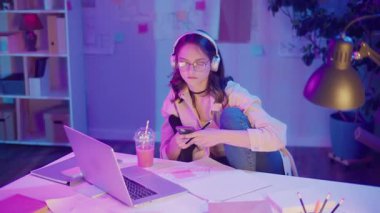 Gen Z Yaşam Tarzı, Dijital Çoklu Görev, Neon Mood. Genç Kız Müzik Dinlerken ve Dizüstü bilgisayar izlerken resim çizer. Neon Işığında Modern Gençliğin Yaratıcı Kaosu 'nda Kişisel İfade