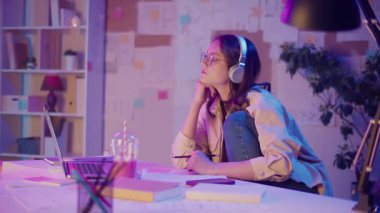 Gen Z Yaşam Tarzı, Dijital Çoklu Görev, Neon Mood. Genç Kız Müzik Dinlerken ve Dizüstü bilgisayar izlerken resim çizer. Neon Işığında Modern Gençliğin Yaratıcı Kaosu 'nda Kişisel İfade