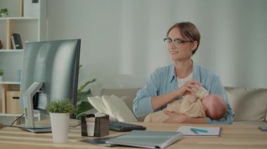 İş hayatı dengesi, melez ebeveynlik, pozitif enerji. Gözlüklü modern bir anne bebeğini şişeyle beslerken ve masasında çalışırken gülümsüyor, kariyer başarısı ve aile bakımını dengeliyor.