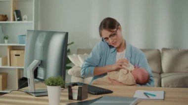 İş hayatı dengesi, melez ebeveynlik, pozitif enerji. Gözlüklü modern bir anne bebeğini şişeyle beslerken ve masasında çalışırken gülümsüyor, kariyer başarısı ve aile bakımını dengeliyor.