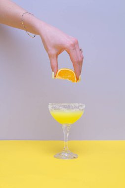 Bardak, içecek ve limon tutan bir kadının eli ile sarı ve gri arka planda minimum kompozisyon