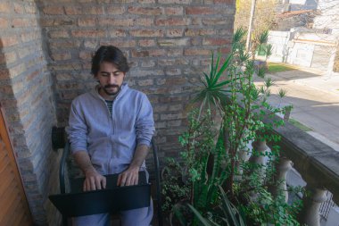 Genç adam evindeki balkonda bitkilerle birlikte dizüstü bilgisayarda çalışıyor.