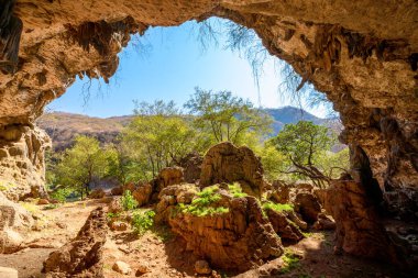 Bir kemer oluşturan doğal mağara girişi, ağaçlı ve engebeli tepeli güneşli, yeşil bir manzaraya doğru bakıyor Zufar, Umman 'da berrak mavi bir gökyüzünün altında, bir keşif hissi veriyor.