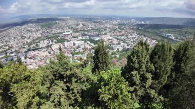 Tiflis 'in verimli yeşillik çerçevelemesi geleneksel ve modern mimarinin bir karışımını ortaya çıkarırken, Kura Nehri güneşli bir gökyüzünün altında canlı şehir manzarası boyunca kıvrılıyor.