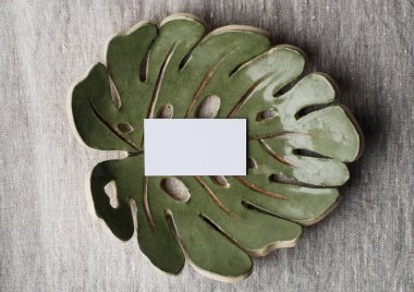 Yaz kırtasiyesi hala hayat dolu. Seramik tabakta Monstera yaprağının boş kartviziti. Gri keten masa örtüsü kumaş arka plan. Marka konsepti. Akdeniz tasarımı. Biçimli düz uzanma