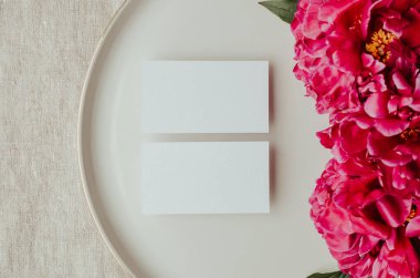 Şakayık çiçekleri ve tomurcuklarla açan dallar. Seramik tabaktaki boş kartvizit modeli. Kadınsı düğün ve kırmızı şakayık çiçekleri. Gri keten masa örtüsü kumaş arka plan. Botanik ve lüks