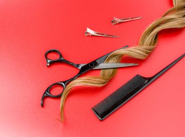 Kırmızı arka planda kıvırcık yapay saç teli olan açık kuaför makasları, profesyonel makas, siyah tarak, metal klipsler ve kesilmiş saç uçları.