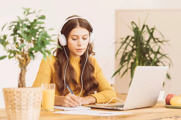 Happy female in wireless headphones studying online course, using pc and writing in notepad