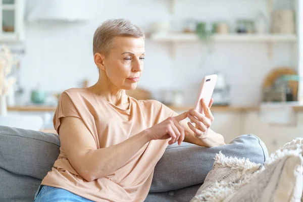 Happy attractive elderly woman chatting with friend on smartphone while sitting on sofa at home