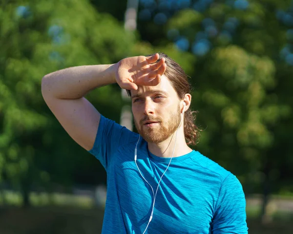 Tired exhausted young sportive man wiping sweat from forehead while exercising outdoors