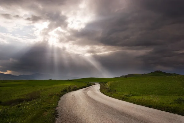 Road crosses prairie covered by clouds into rays of sun — Stock Photo, Image