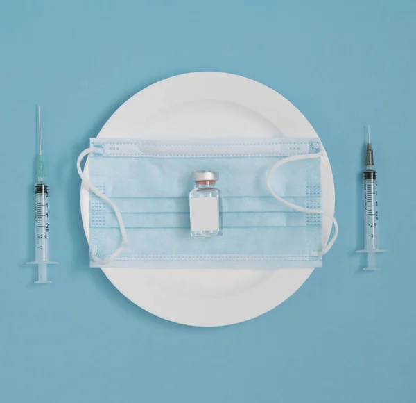 The vial and face mask lay down on a plate, with two syringes by the side. Minimal flat lay pandemic concept on pastel blue background