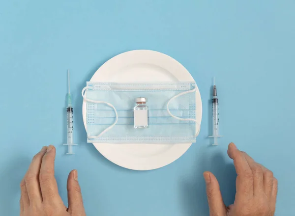 The vial and face mask lay down on a plate, with two syringes and hands by the side. Minimal flat lay pandemic concept on pastel blue background