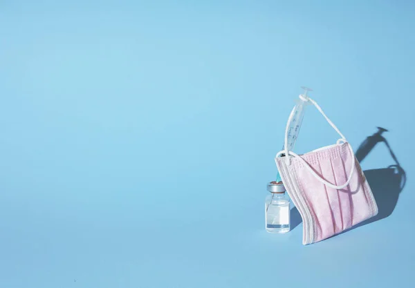 Face mask hanging on syringe and vial on pastell blue background, minimal abstract pandemic vaccination concept