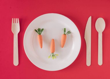 Tahta bir kaşık, çatal ve bıçakla beyaz bir tabakta suni havuç kırmızı bir arka plan masasına uzanıyordu.