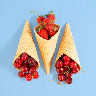 Olgun çilek ve kirazla dolu dondurma külahları mavi arka planda uzanıyorlar. Kare düz kompozisyon. Minimum yaz tazeleyici kavram