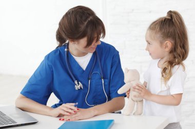 Genç, gülümseyen kadın doktor ve oyuncak ayıyla küçük hastası.