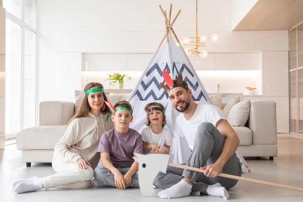 Happy family of parents and two children playing indian at home, wigwam tent, toy horse, feather roach