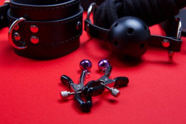 Ağız ve kelepçenin arkasında meme ucu klipsi var. Kırmızı arka planda, ceza, itaat, seks ve tutku için bdsm oyuncakları.